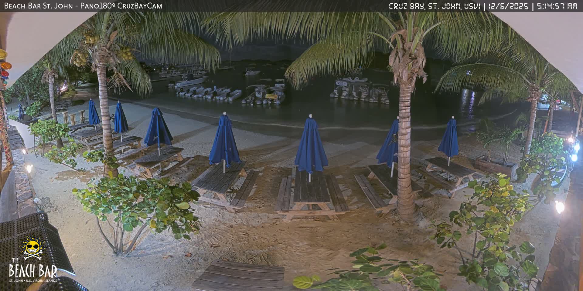 A serene nighttime view captures a sandy beach with several picnic tables and closed blue umbrellas surrounded by palm trees and lush foliage, facing a calm bay dotted with numerous anchored boats under clear, calm weather.