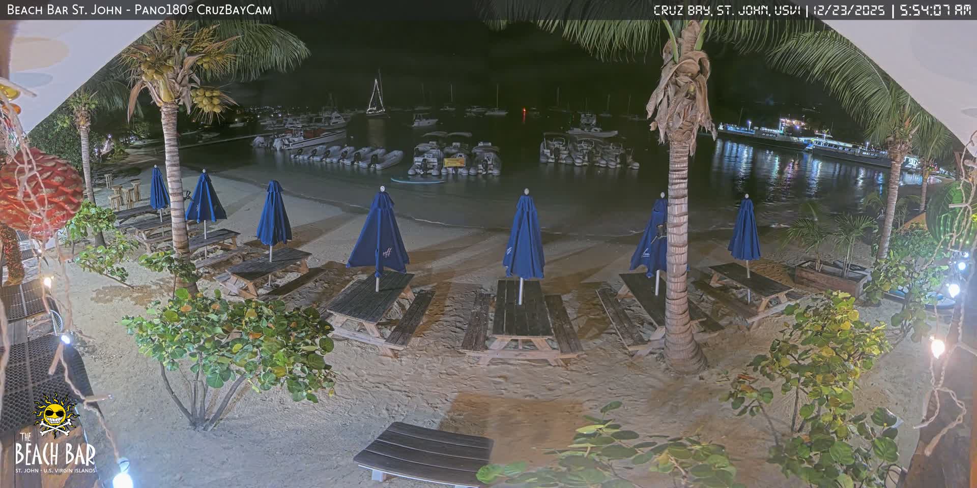 Under a clear, dark sky in the early morning, a tropical sandy beach features numerous wooden picnic tables, folded blue umbrellas, and palm trees, overlooking a calm bay bustling with anchored boats and their reflections.