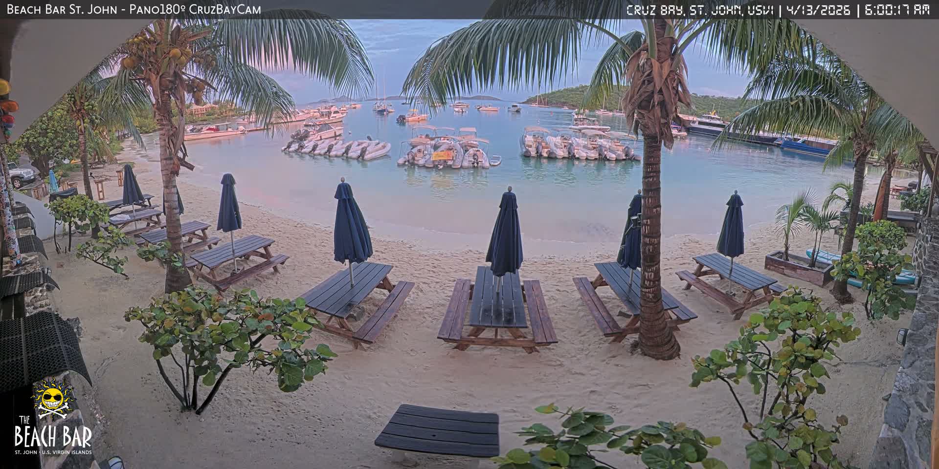 Under a clear, dark sky in the early morning, a tropical sandy beach features numerous wooden picnic tables, folded blue umbrellas, and palm trees, overlooking a calm bay bustling with anchored boats and their reflections.