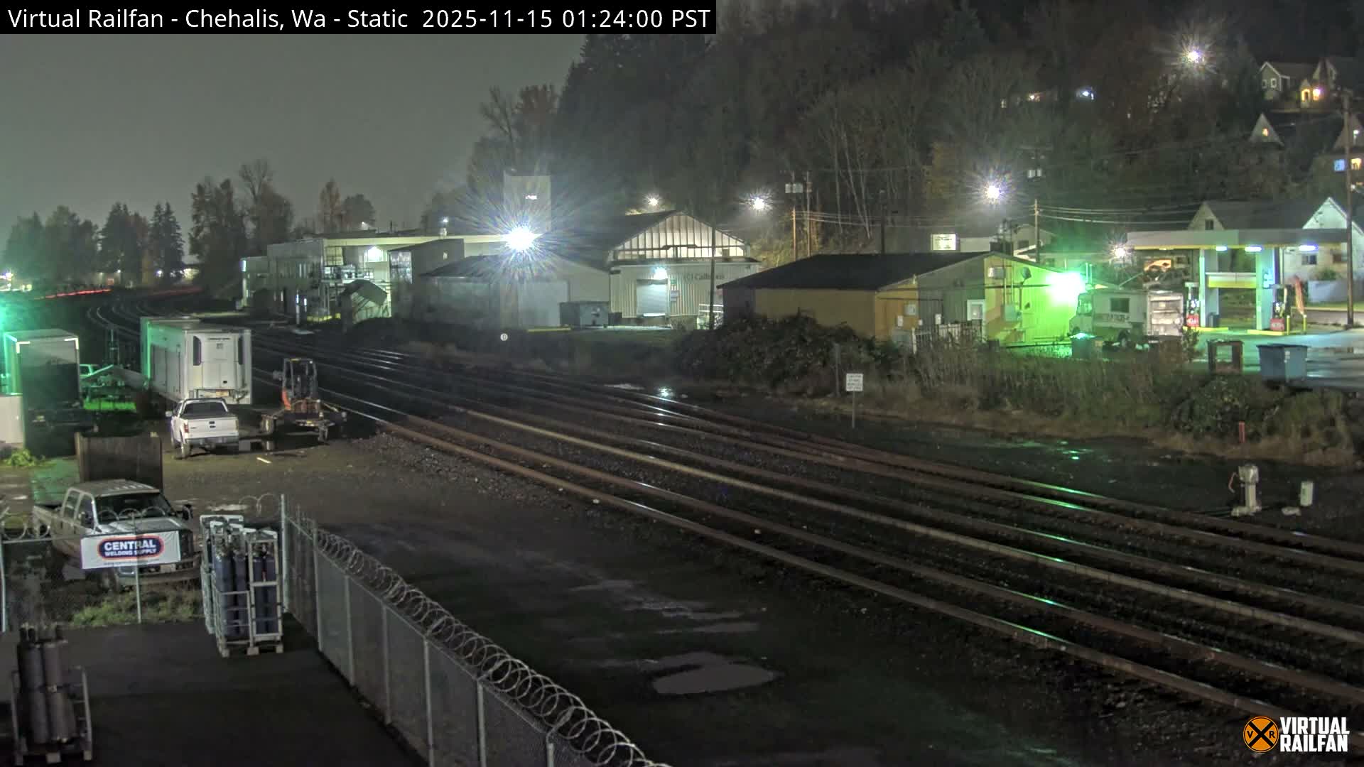 A nighttime scene captures a rail yard with numerous tracks, brightly lit industrial buildings, and parked vehicles including trucks and an excavator, under a damp, overcast sky.