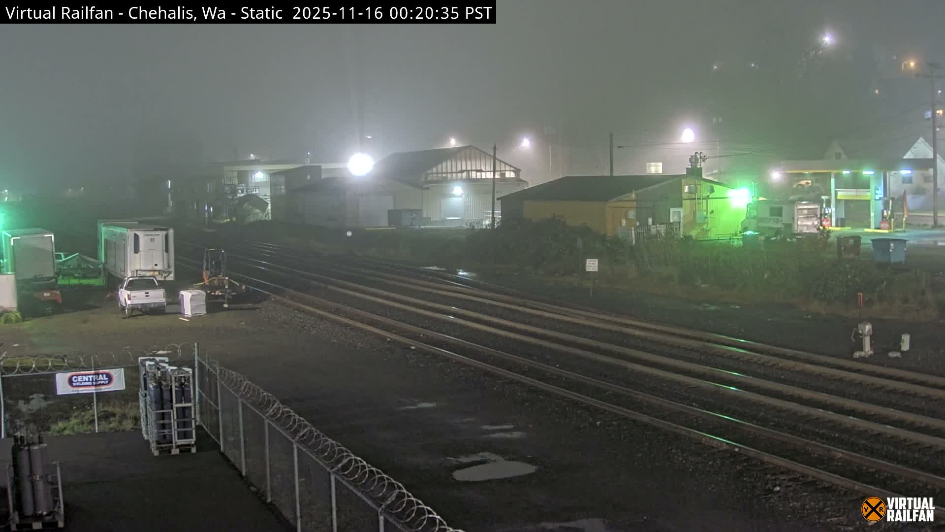 A foggy night scene reveals multiple parallel railroad tracks stretching into the distance, bordered by industrial buildings and a gas station illuminated by artificial lights, with wet ground and parked vehicles in the foreground.