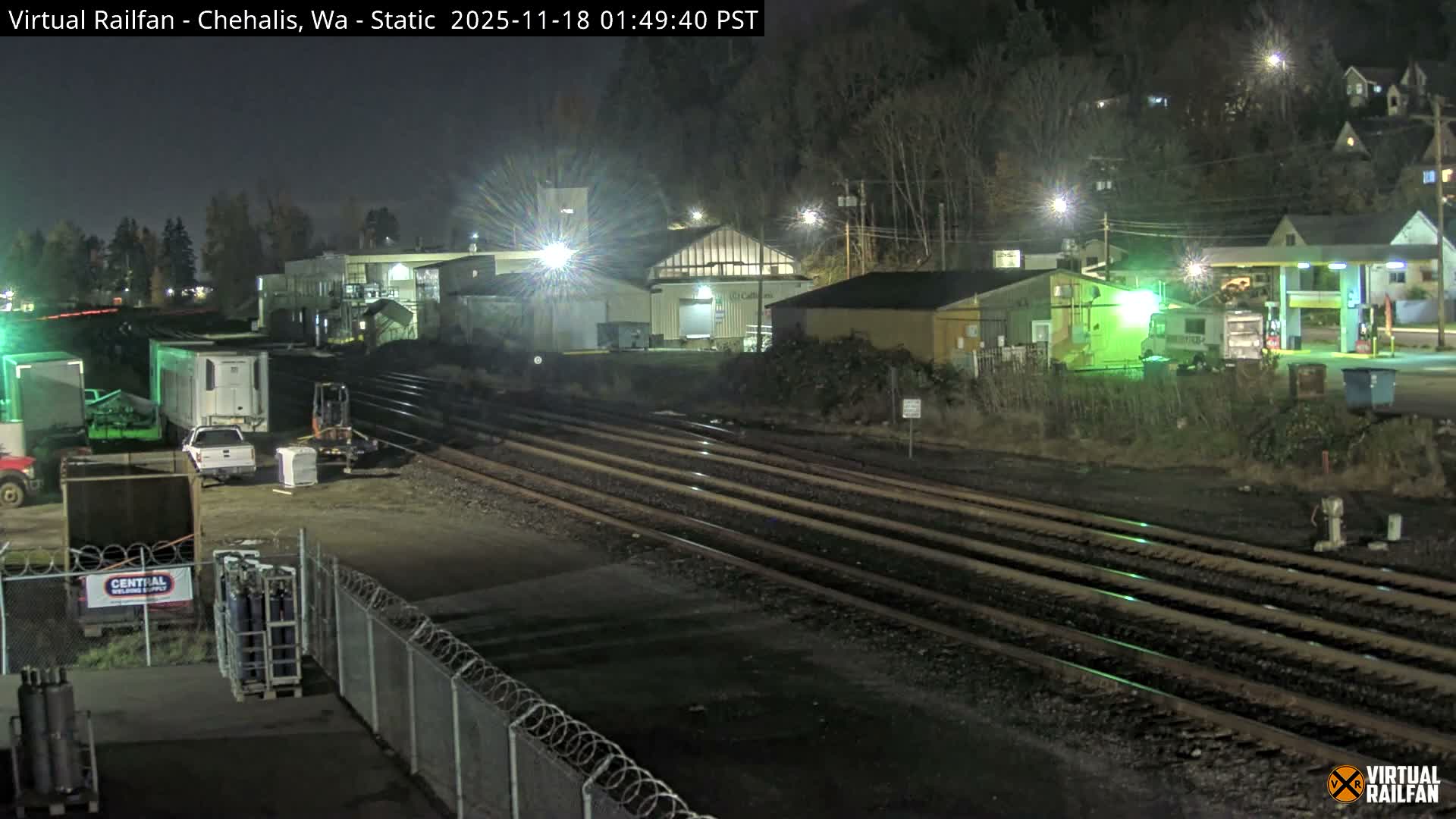 A foggy night scene reveals multiple parallel railroad tracks stretching into the distance, bordered by industrial buildings and a gas station illuminated by artificial lights, with wet ground and parked vehicles in the foreground.