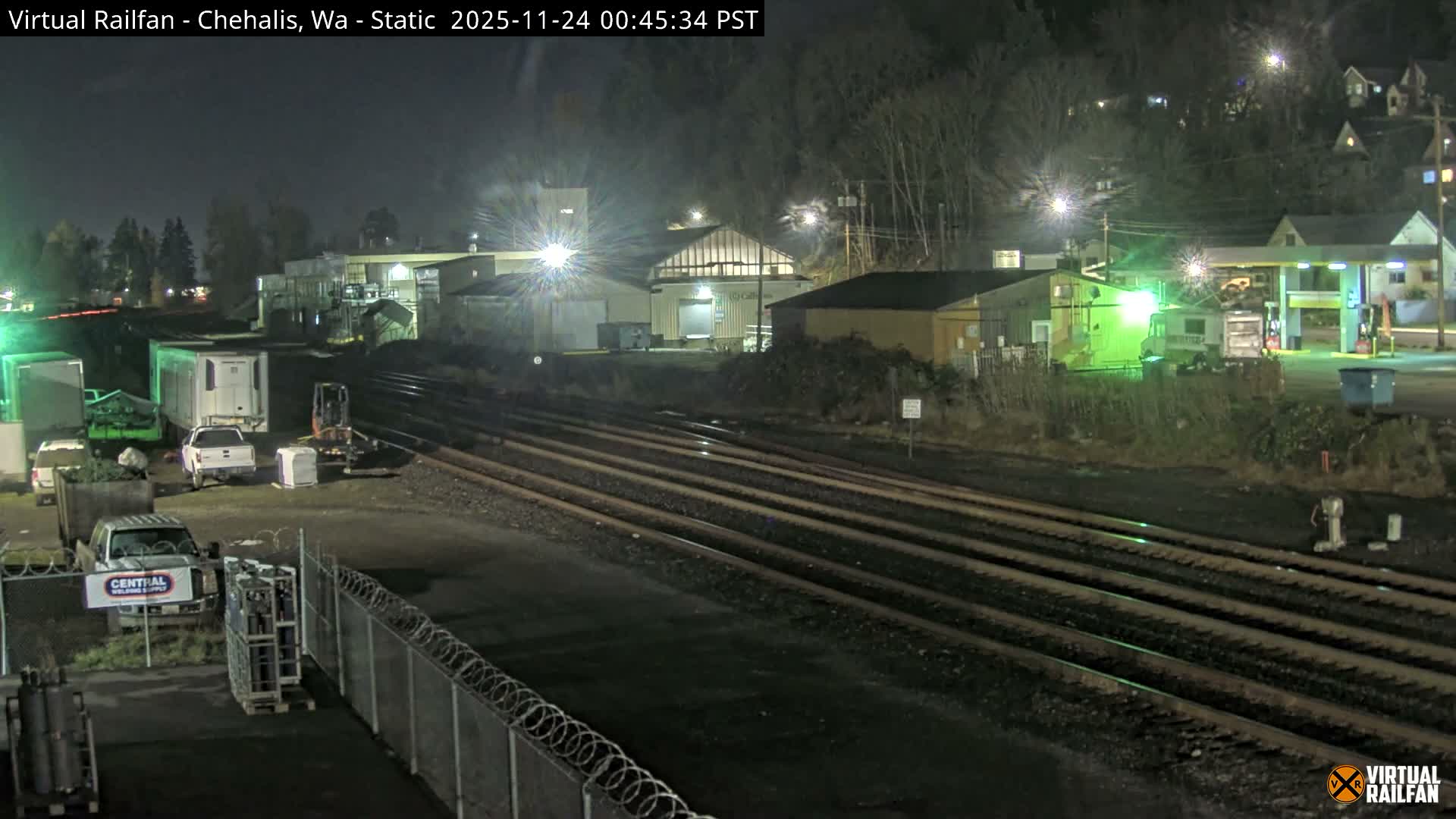 Multiple railroad tracks stretch through an illuminated industrial and commercial area with various buildings, parked vehicles, and a gas station, all under a clear night sky.