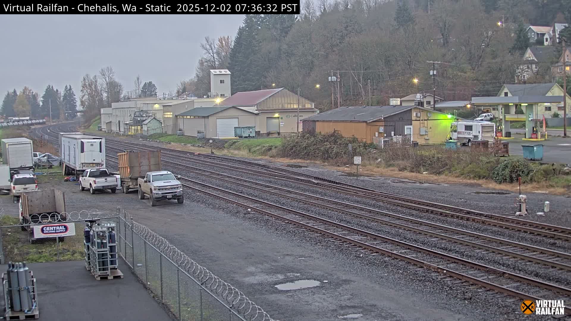 On an overcast morning, an industrial rail yard features multiple train tracks, various buildings, and several parked vehicles, with puddles on the ground and tree-covered hills in the background.