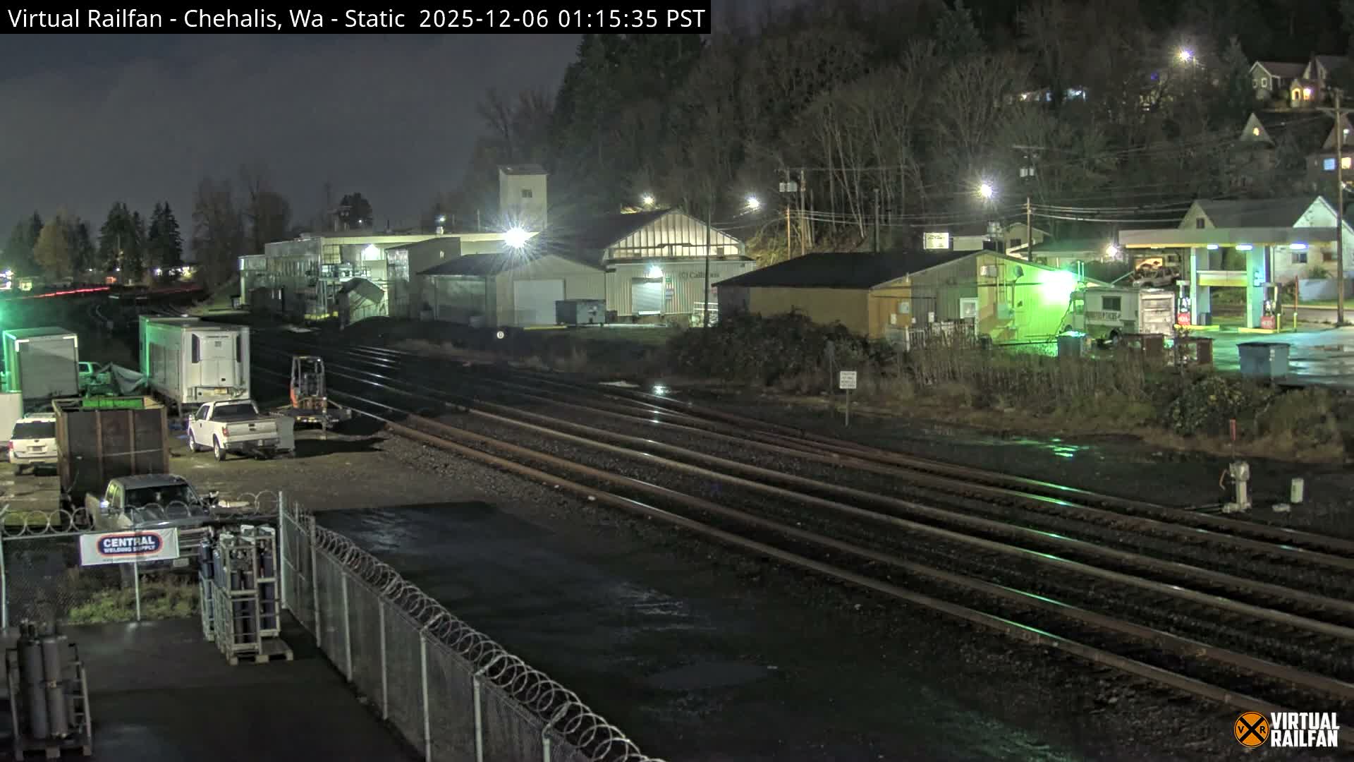 Multiple wet railroad tracks traverse an illuminated industrial and commercial district at night, featuring various buildings, parked vehicles, and a distant gas station under an overcast sky.
