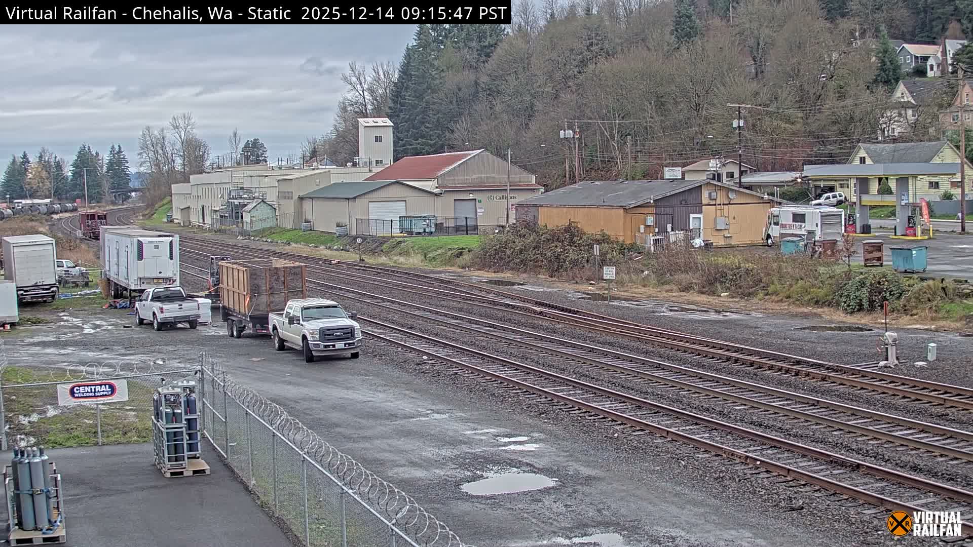 Multiple wet railroad tracks traverse an illuminated industrial and commercial district at night, featuring various buildings, parked vehicles, and a distant gas station under an overcast sky.