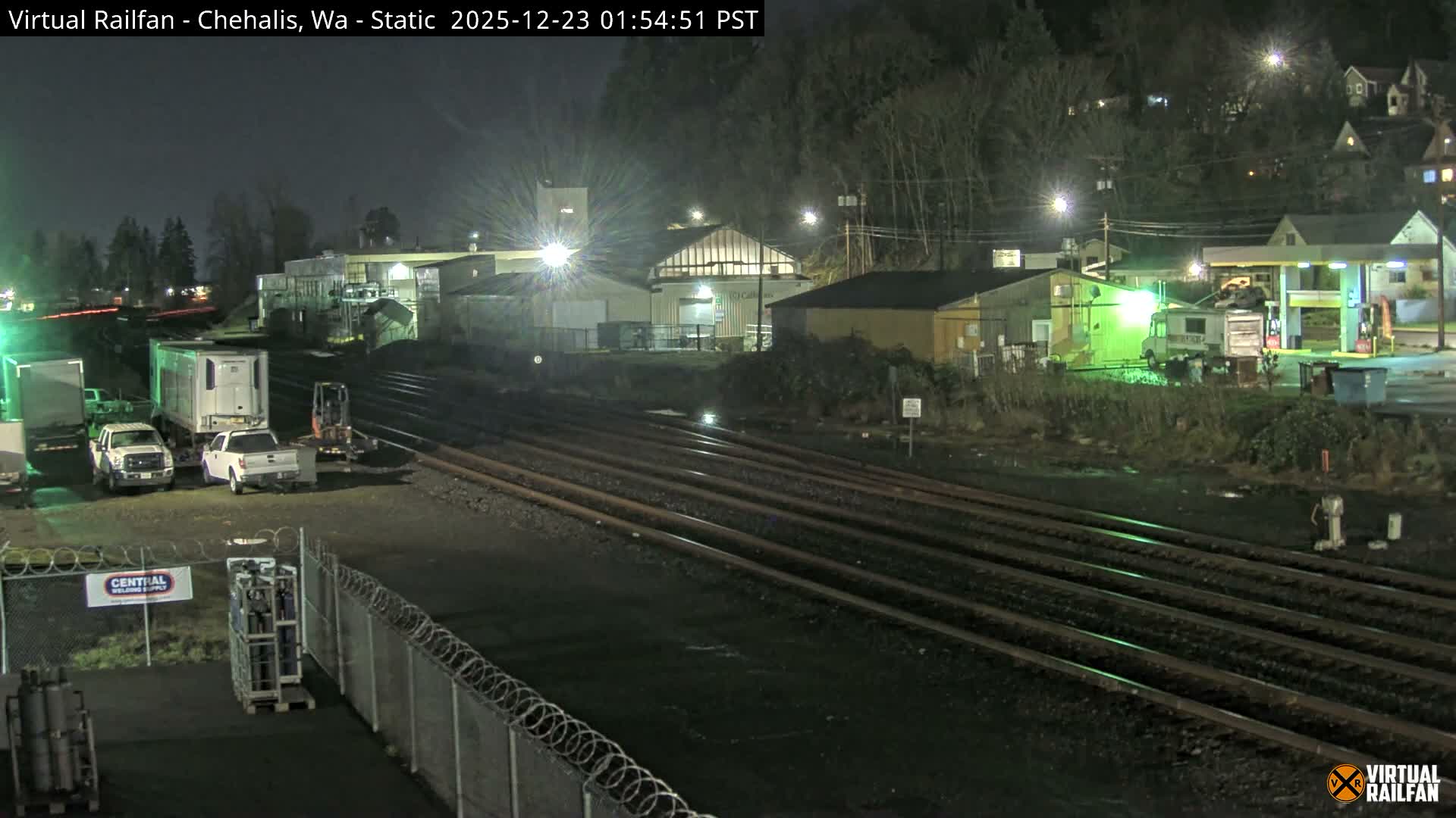 At night, multiple train tracks traverse a damp industrial area featuring various buildings, parked vehicles, and a brightly lit gas station, with a dark, wooded hillside dotted with illuminated houses in the background.