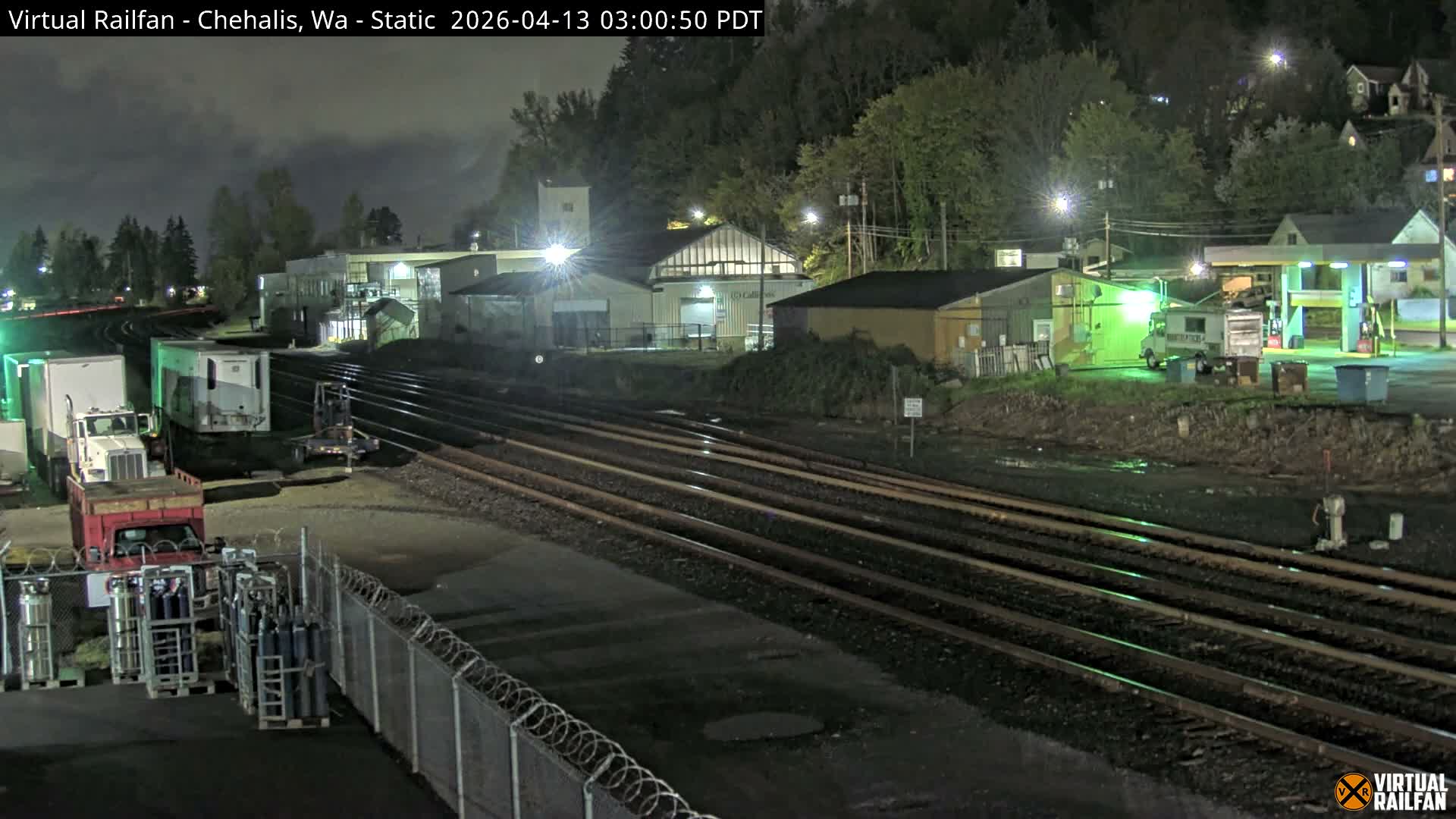 At night, multiple train tracks traverse a damp industrial area featuring various buildings, parked vehicles, and a brightly lit gas station, with a dark, wooded hillside dotted with illuminated houses in the background.