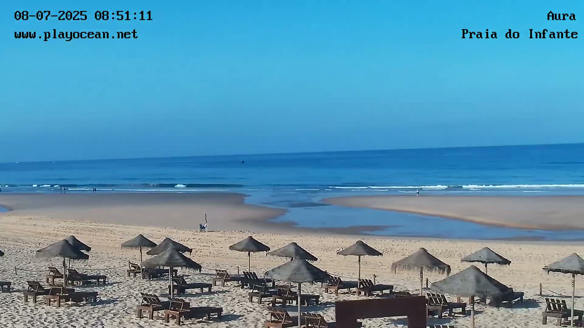 A sandy beach with numerous thatched umbrellas and lounge chairs is shown, overlooking a calm ocean under a clear blue sky.