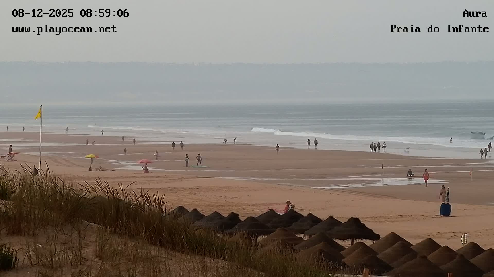 A hazy beach scene shows people walking and swimming in the ocean, with several umbrellas and beach huts visible on the sand.