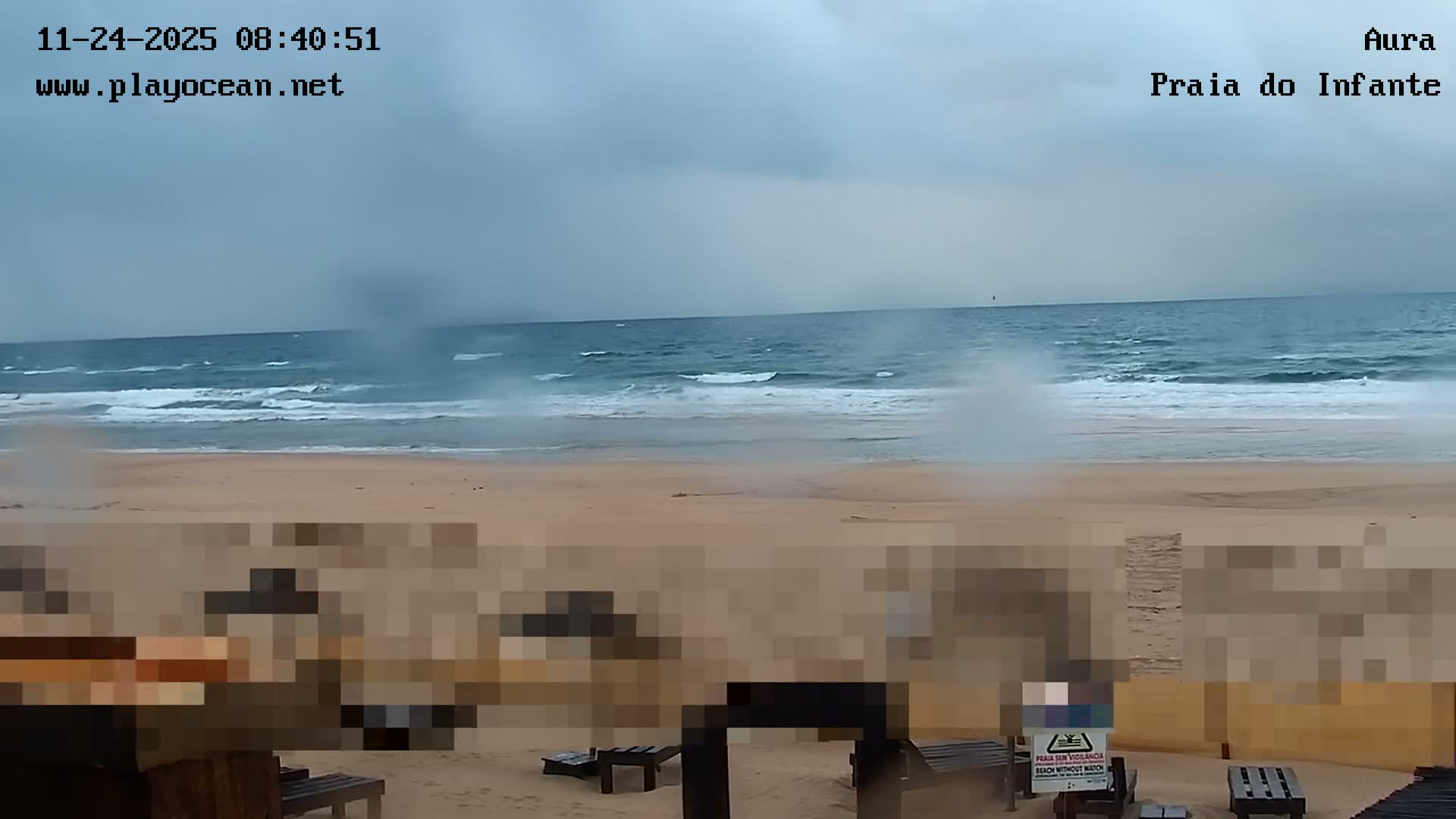 A sandy beach stretches towards a choppy, blue-grey ocean with breaking waves under a heavily overcast and possibly rainy sky.