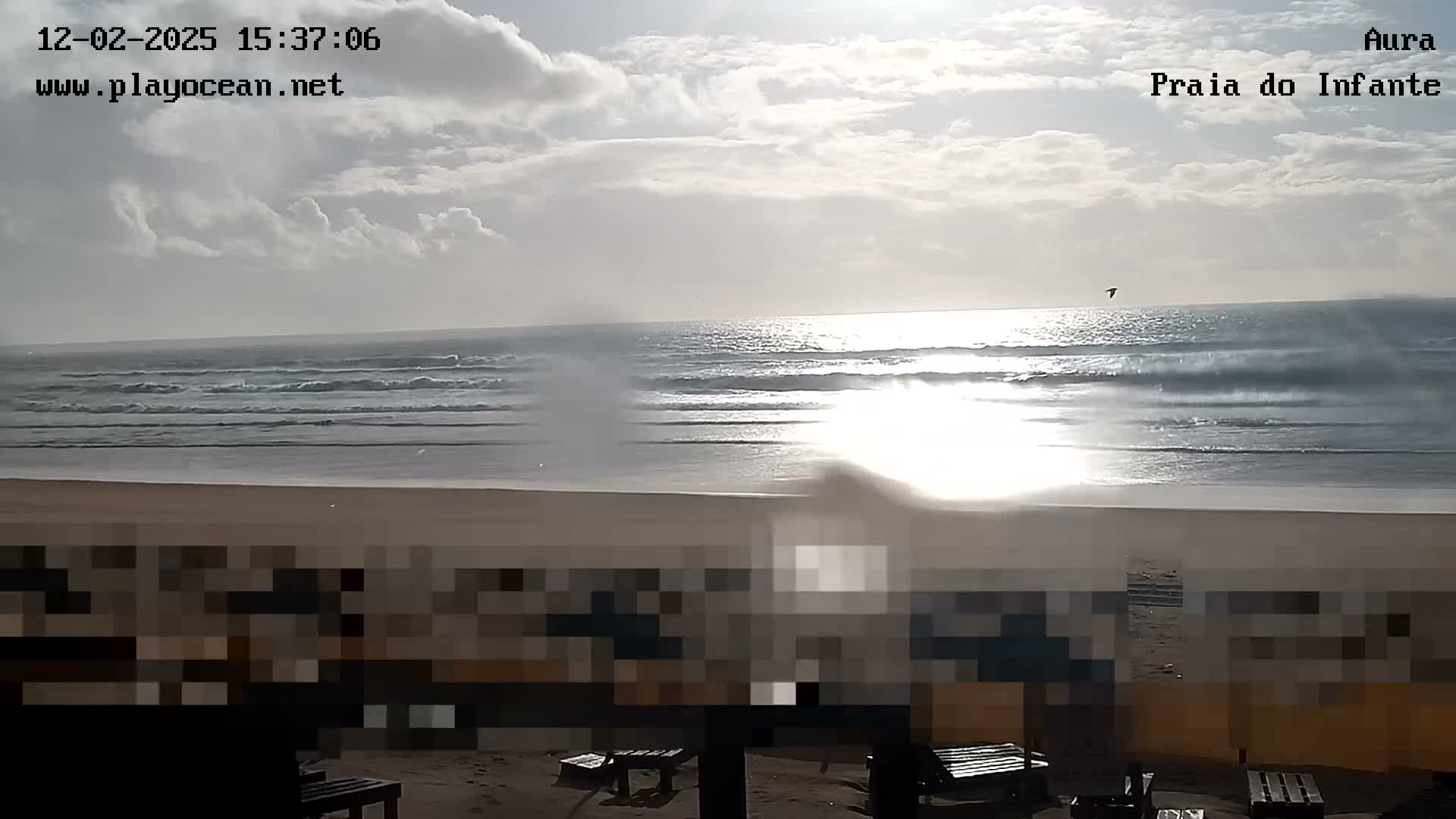 A bright and partly cloudy day reveals a wide sandy beach beside an ocean with rolling waves and a strong sun glare, under a sky scattered with clouds where a single bird flies.
