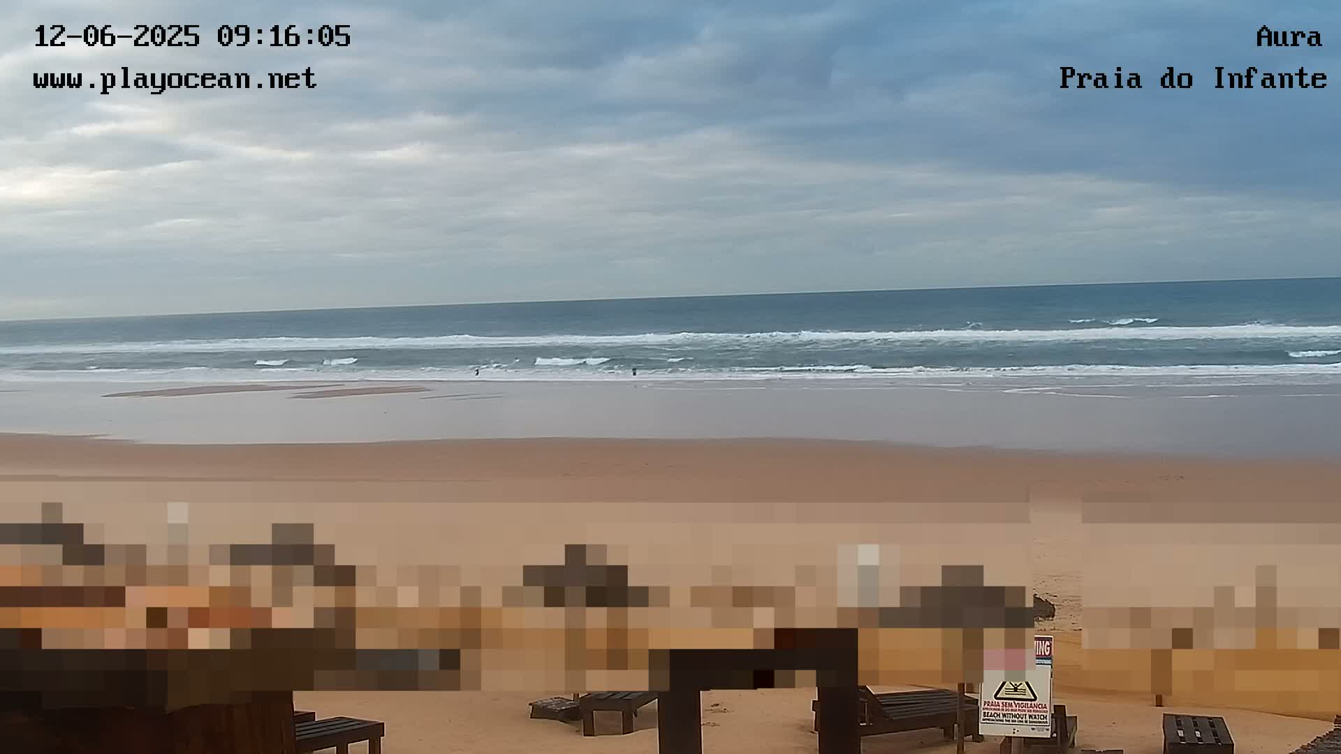 A cloudy day overlooks a sandy beach with breaking waves and a few individuals in the water, framed by indistinct, pixelated structures in the foreground.