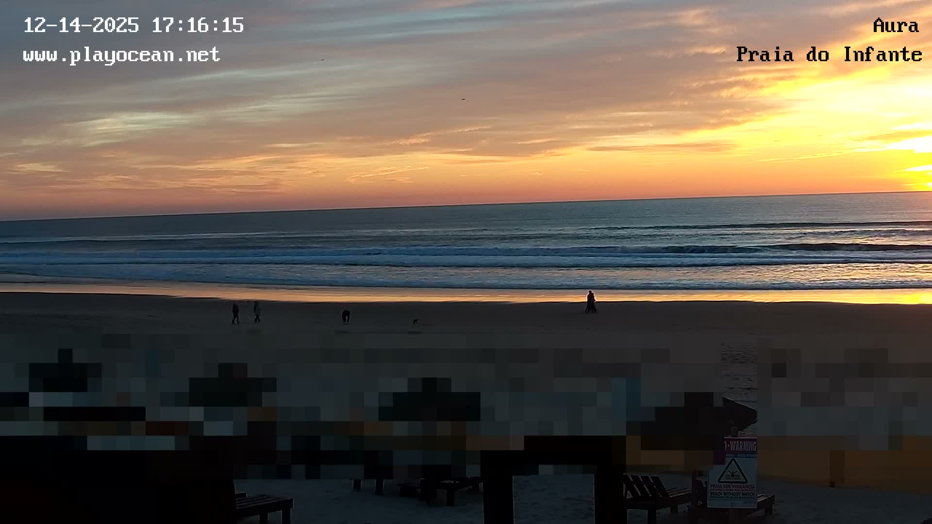 A cloudy day overlooks a sandy beach with breaking waves and a few individuals in the water, framed by indistinct, pixelated structures in the foreground.