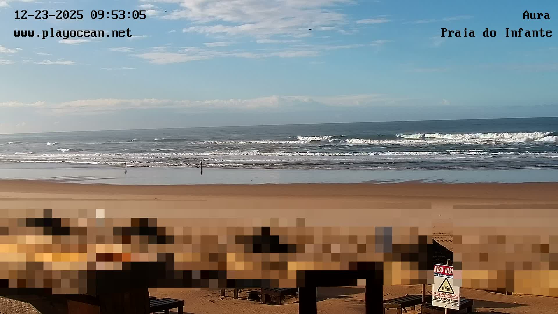 The image shows a wide sandy beach with several people standing near the ocean's edge as waves crash, all under a bright, partly cloudy sky.