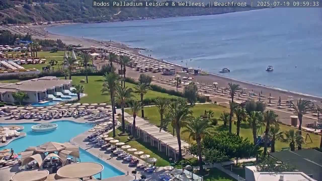 An aerial view shows a resort with a swimming pool and beach umbrellas, adjacent to a sandy beach with more umbrellas and calm ocean water under a sunny sky.