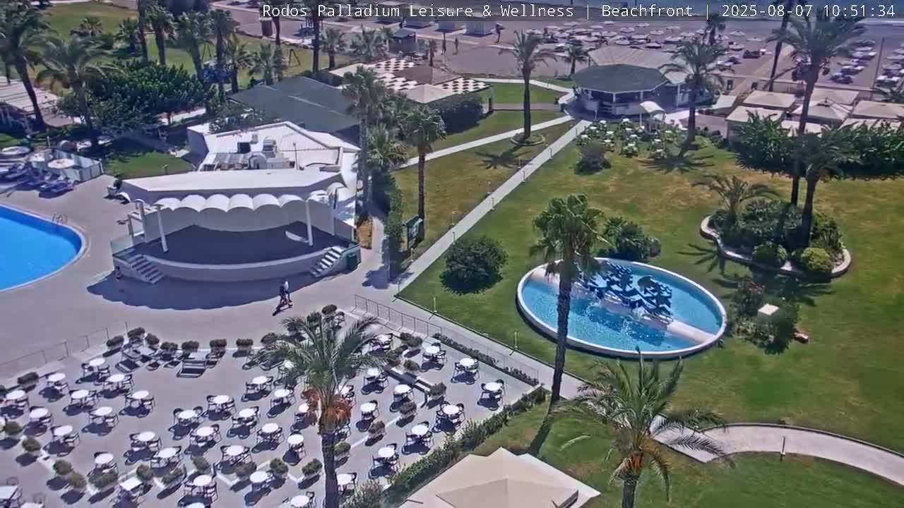 An aerial view shows a resort area with a swimming pool, a stage, several seating areas with tables and chairs, a small circular pool with a fountain, and palm trees under a sunny sky.