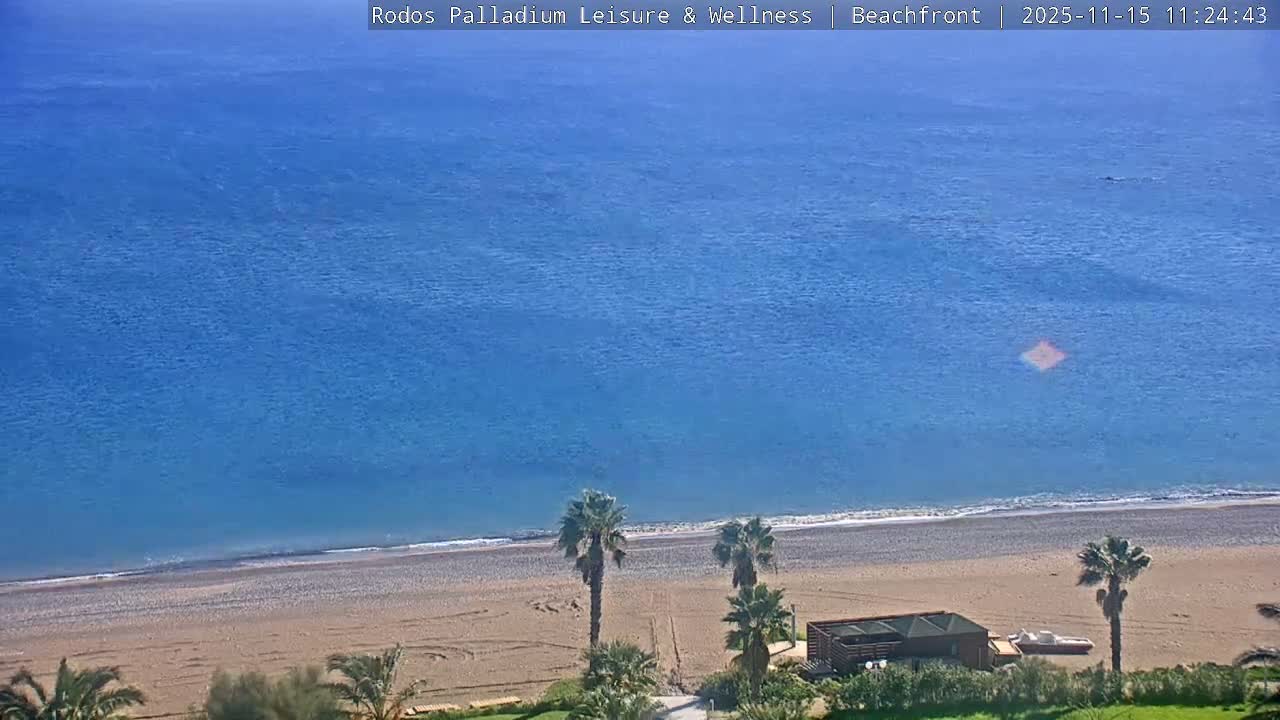 A calm, deep blue sea meets a light brown pebble beach under sunny conditions, featuring several palm trees and a small building with a boat nearby.