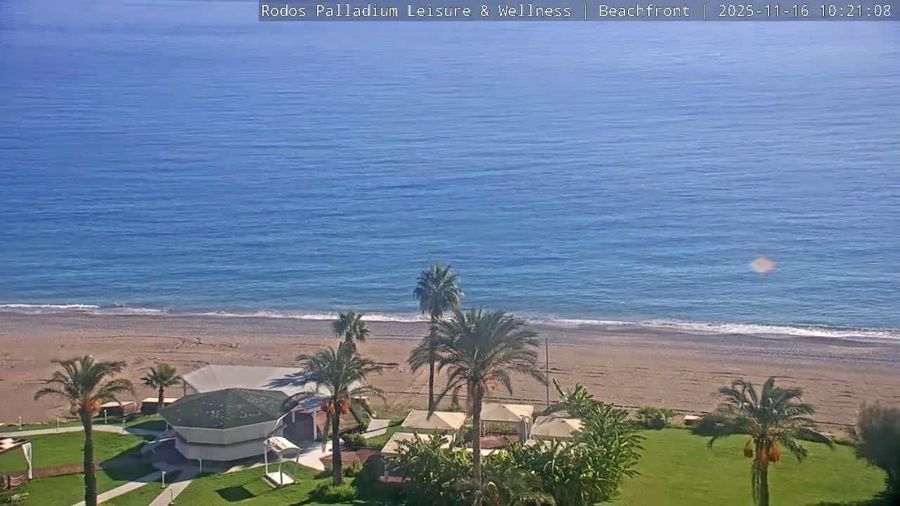 The image shows a bright, sunny beachfront scene with a calm blue sea, a wide sandy beach, and lush green grounds featuring palm trees and resort-like structures.