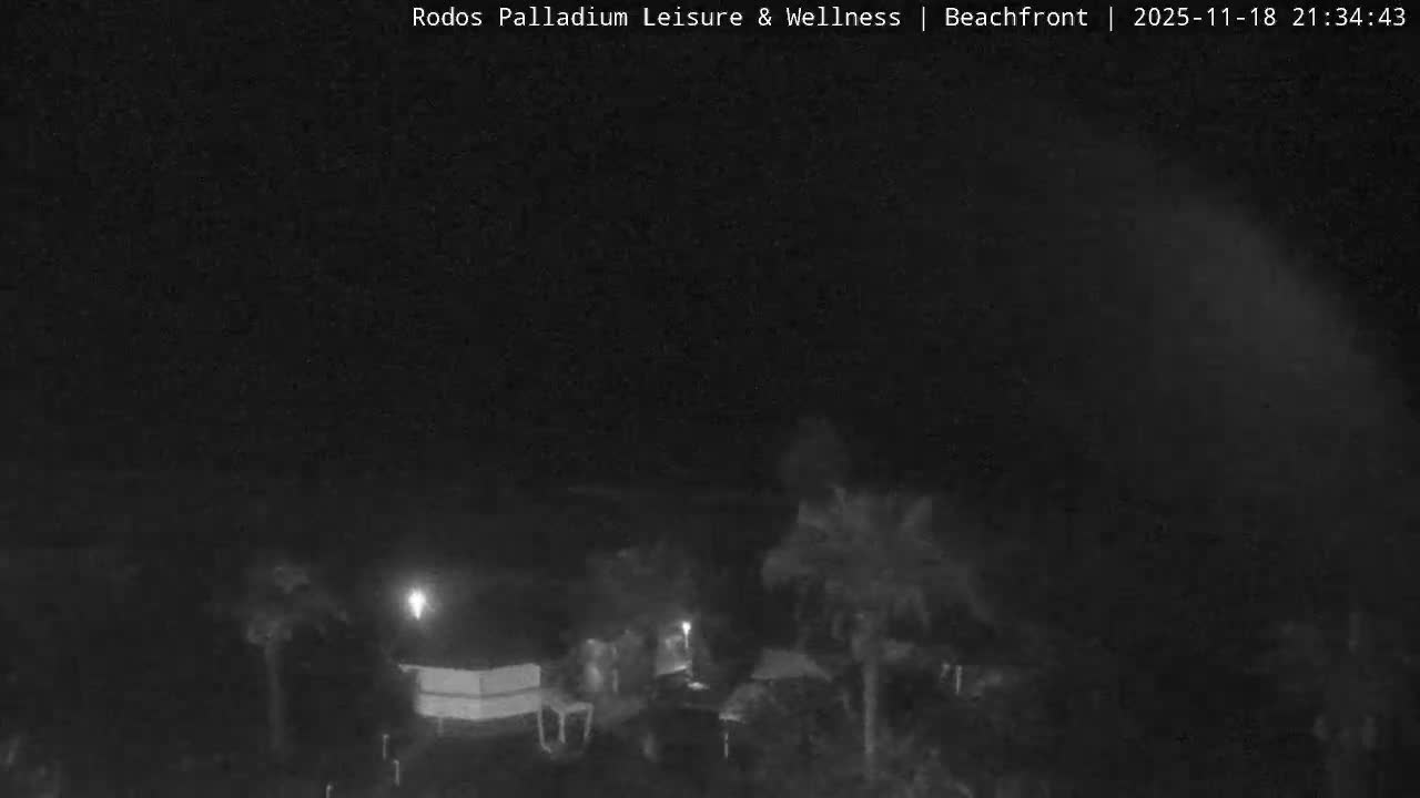 The image shows a bright, sunny beachfront scene with a calm blue sea, a wide sandy beach, and lush green grounds featuring palm trees and resort-like structures.