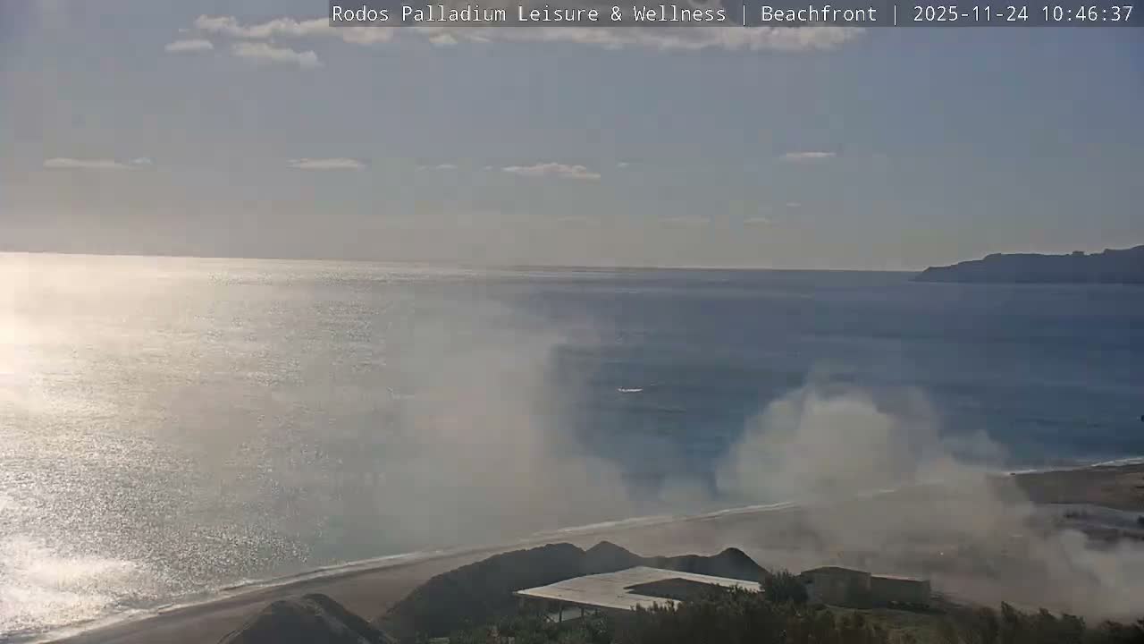On a bright, partly cloudy day, large plumes of smoke or mist rise from a beach and coastal area, bordering a shimmering sea where a small boat is visible, with distant land on the horizon.