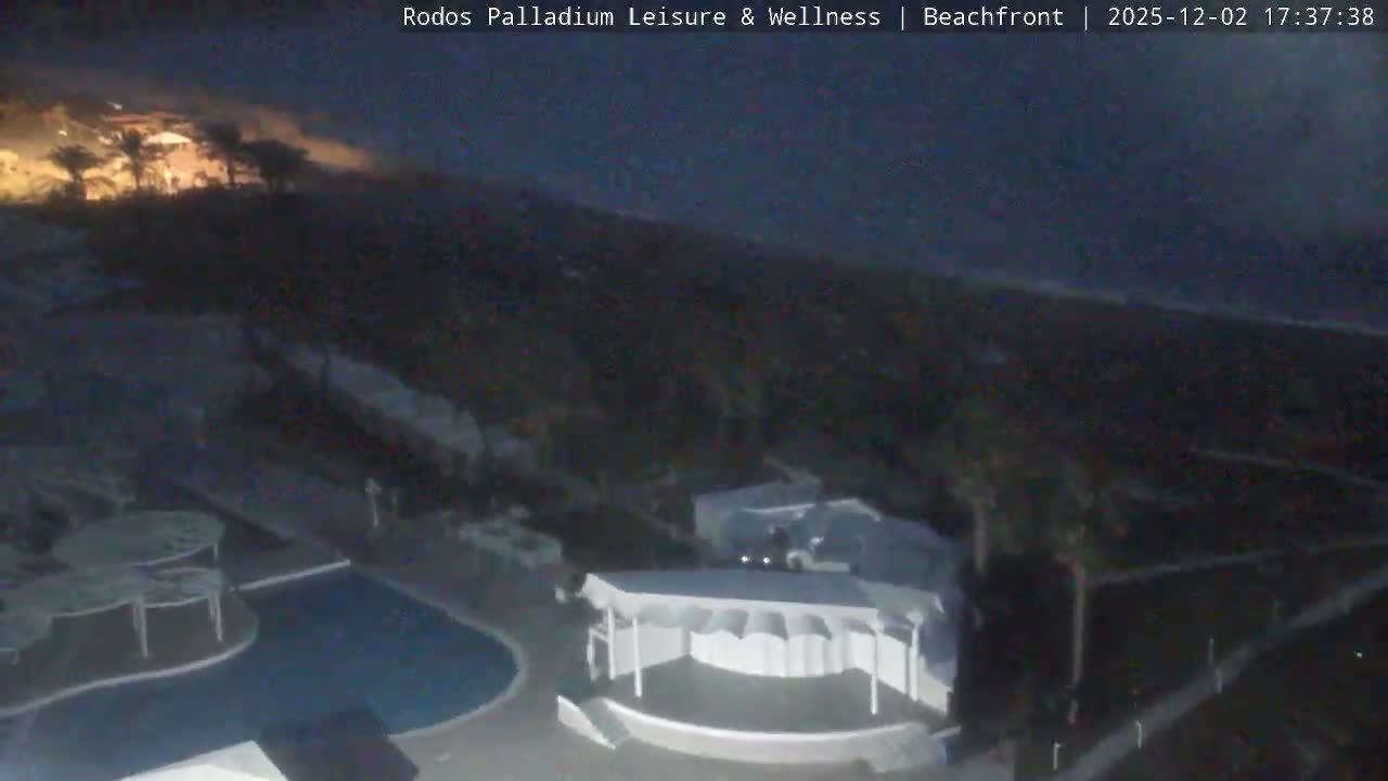 A dark, high-angle view captures a beach resort at night under clear skies, revealing a swimming pool, an outdoor stage, and palm trees, with the sea dimly visible in the background.