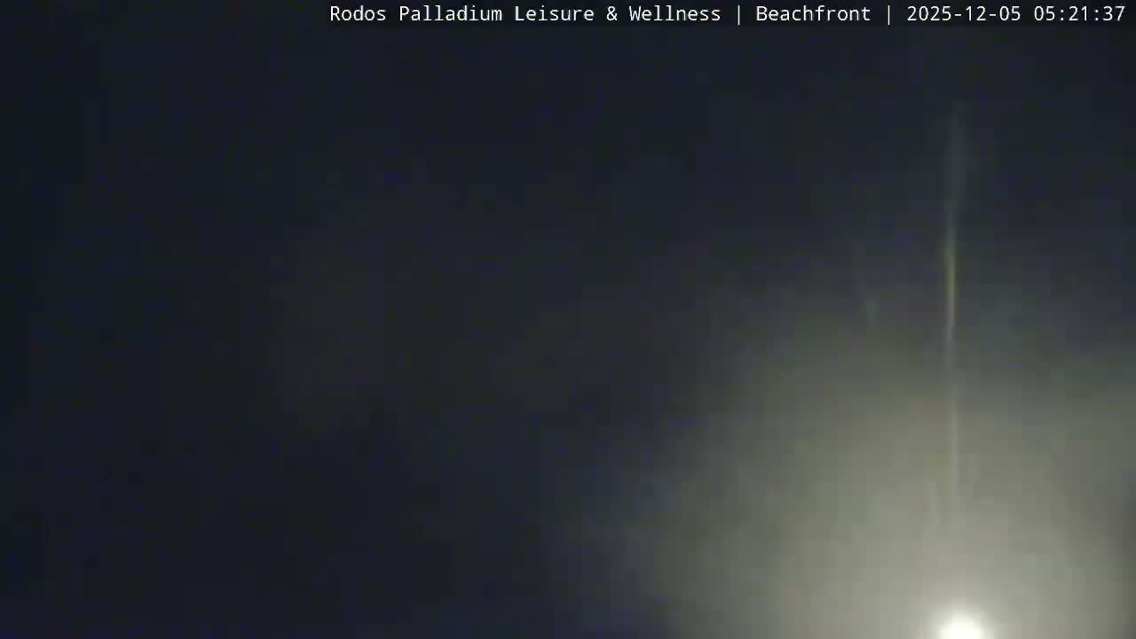 A dark, high-angle view captures a beach resort at night under clear skies, revealing a swimming pool, an outdoor stage, and palm trees, with the sea dimly visible in the background.