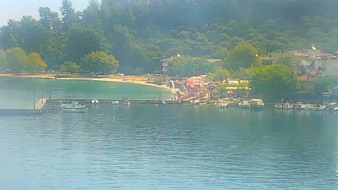 A hazy view of a calm bay shows a small beach with umbrellas and people, a few boats moored at a dock, and buildings nestled among green hills.