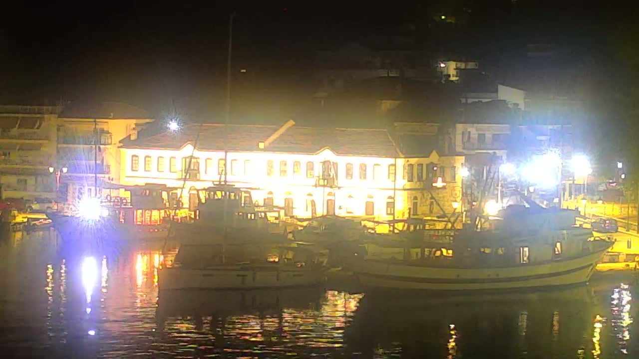 A hazy view of a calm bay shows a small beach with umbrellas and people, a few boats moored at a dock, and buildings nestled among green hills.