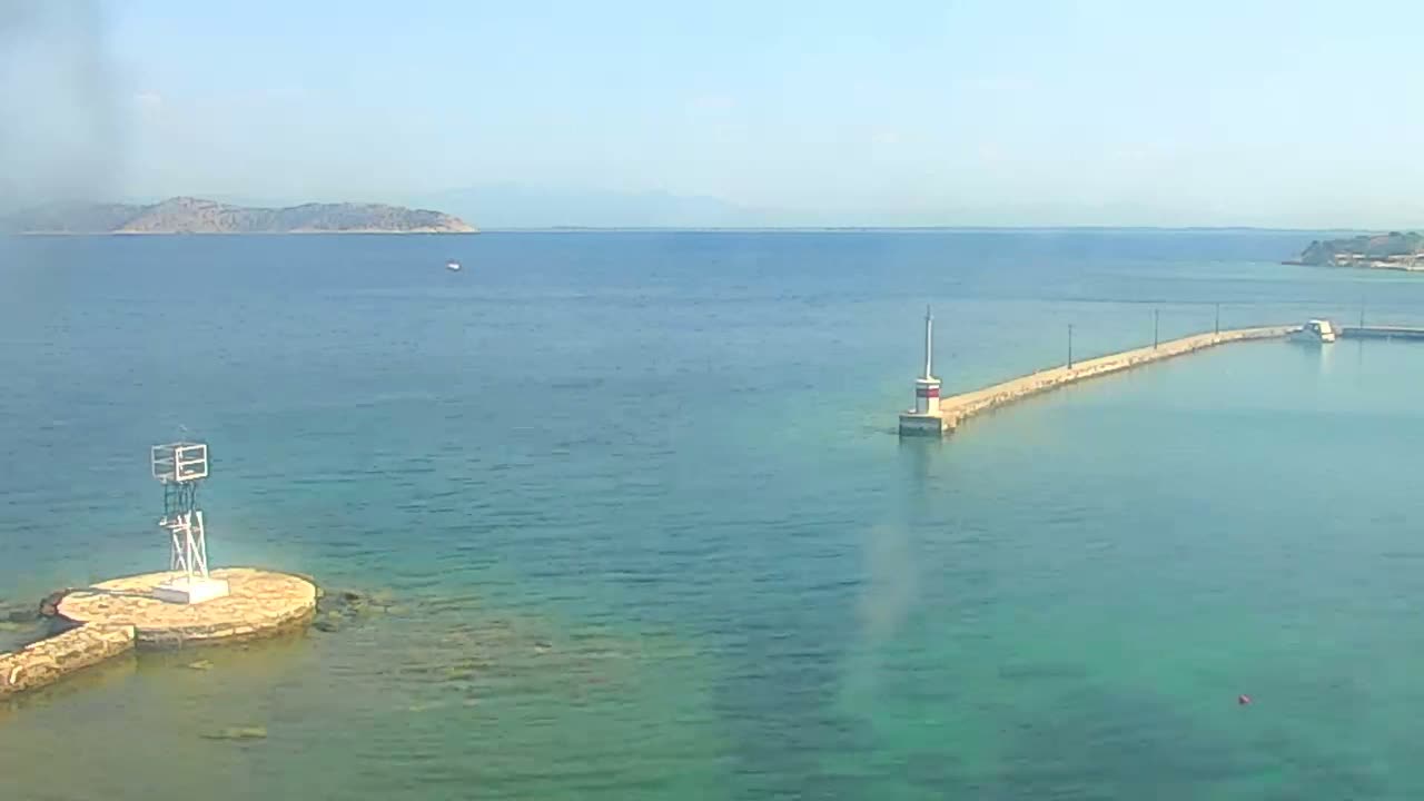 A long jetty extends into a calm, clear turquoise sea under a bright, sunny sky, with a small island visible in the distance.