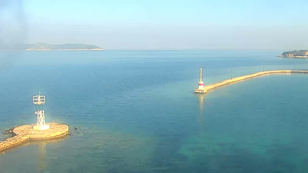 Under a bright, clear sky, a tranquil turquoise sea is dotted with two navigational beacons on stone structures, one a white lattice frame and the other a red and white striped lighthouse at the end of a long, curved pier, with distant islands visible on the horizon.