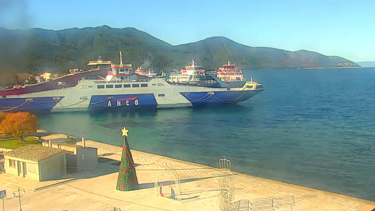Two large ferries are docked in a serene harbor, flanked by mountains, under a clear, sunny sky, with a festive Christmas tree and a small building visible on the paved waterfront.