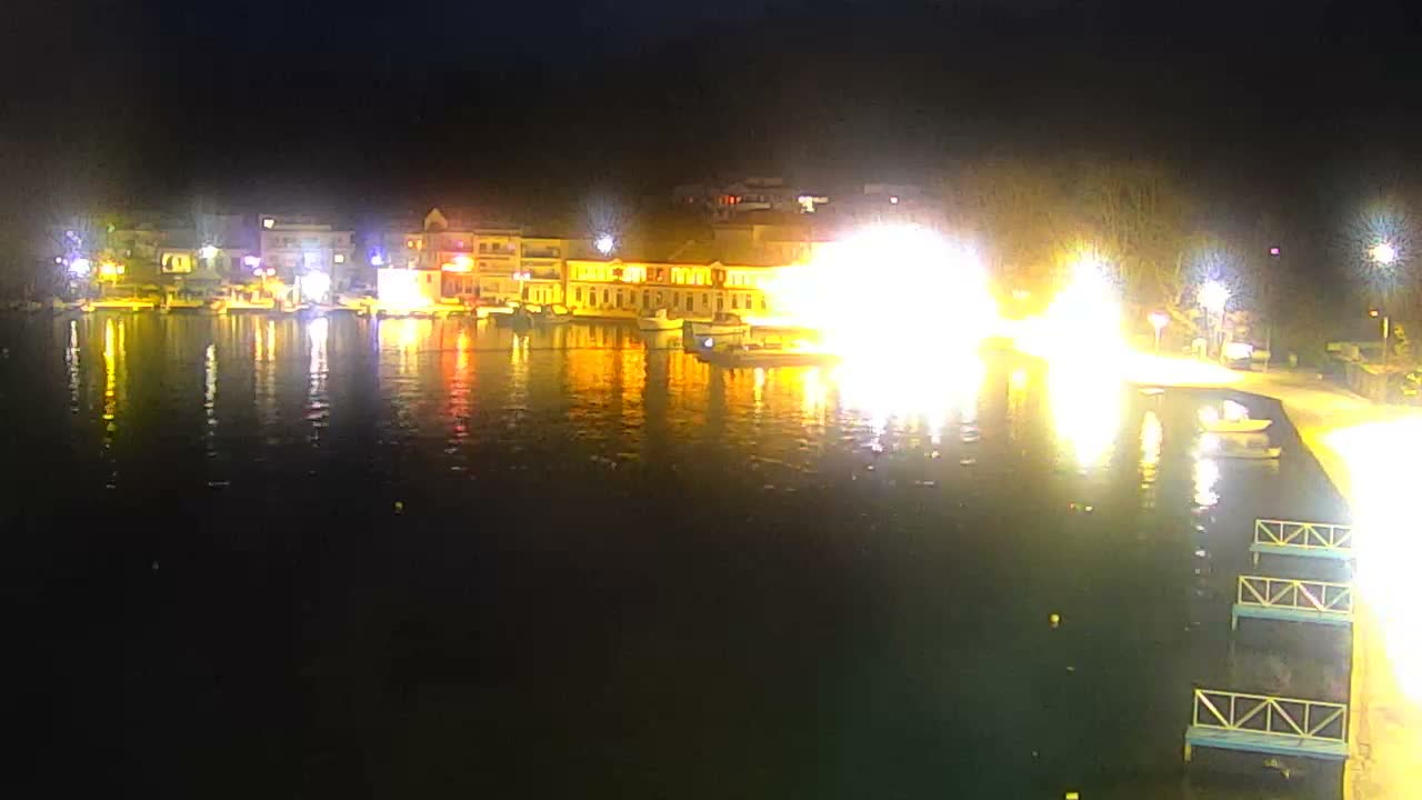 The image shows a nighttime view of a brightly lit waterfront town featuring buildings, several boats, and a path alongside the dark, reflective water, with an intensely bright light source dominating the right side, under clear weather conditions.
