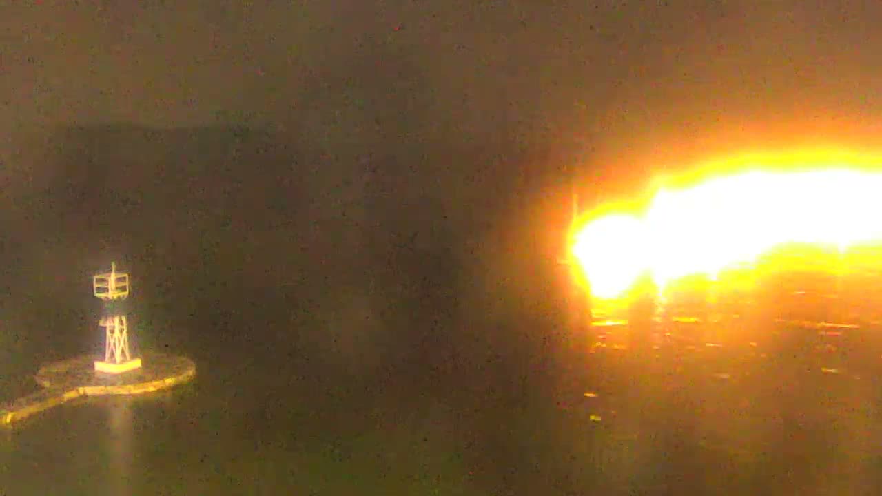 The image shows a nighttime view of a brightly lit waterfront town featuring buildings, several boats, and a path alongside the dark, reflective water, with an intensely bright light source dominating the right side, under clear weather conditions.