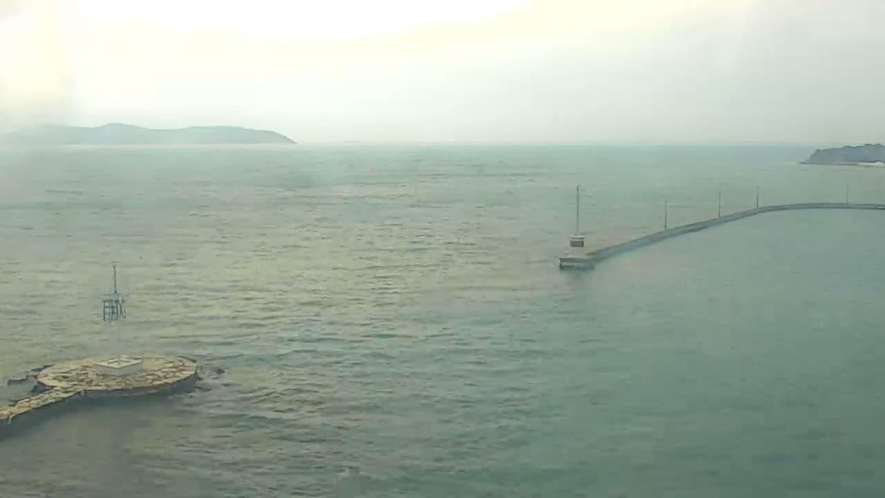 Under a hazy, overcast sky, a calm greenish sea is seen with a circular concrete platform and pole in the foreground, and a long, curved pier with streetlights extending to a small building in the mid-ground, all framed by faint distant land.