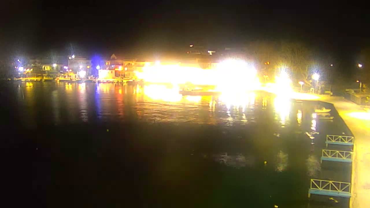 Under a hazy, overcast sky, a calm greenish sea is seen with a circular concrete platform and pole in the foreground, and a long, curved pier with streetlights extending to a small building in the mid-ground, all framed by faint distant land.