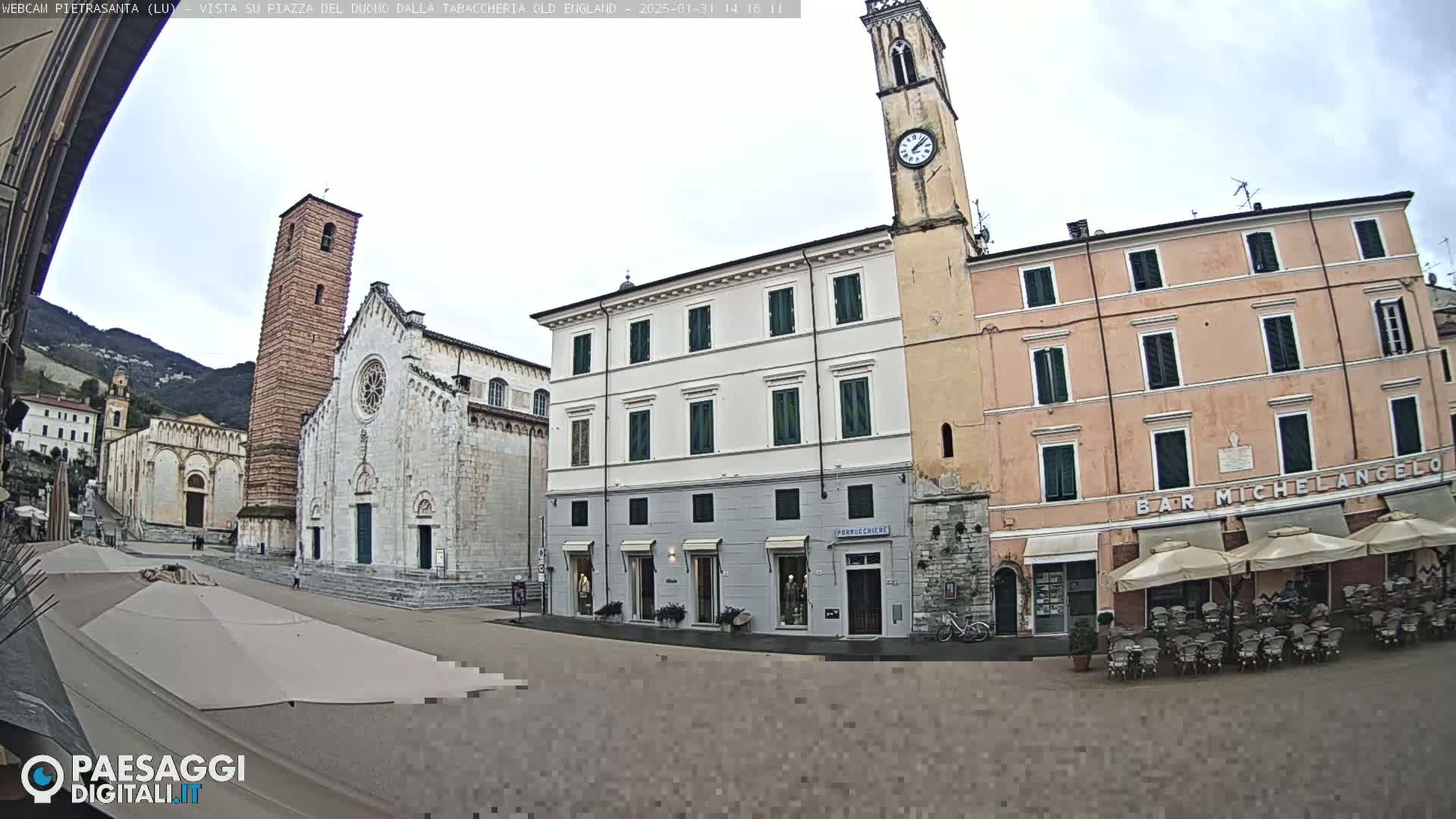 A town square is viewed from a slightly elevated perspective, showing several buildings including a church with a tall bell tower, and an outdoor café under umbrellas on an overcast day.