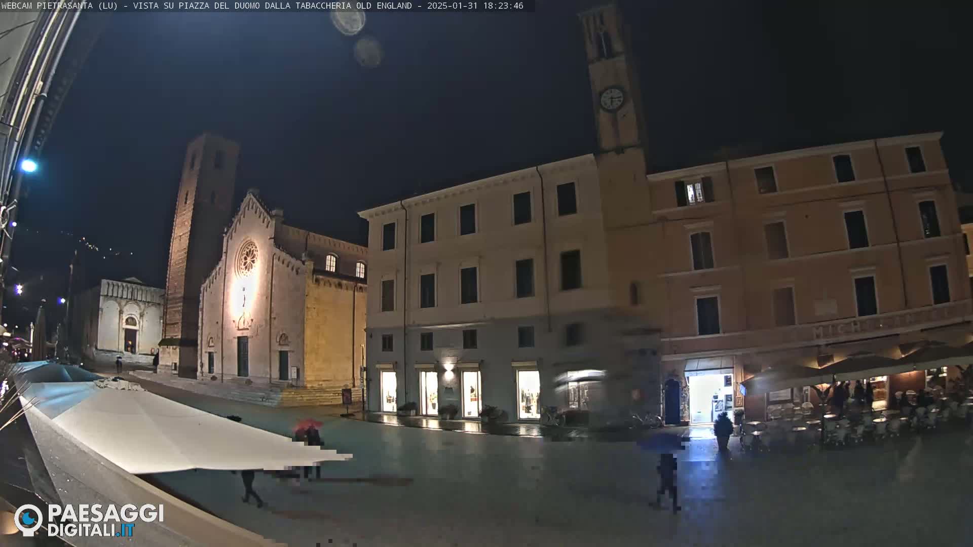 A rain-slicked town square at night, illuminated by streetlights, features a church with a tall bell tower and several buildings, with a few people walking under umbrellas.