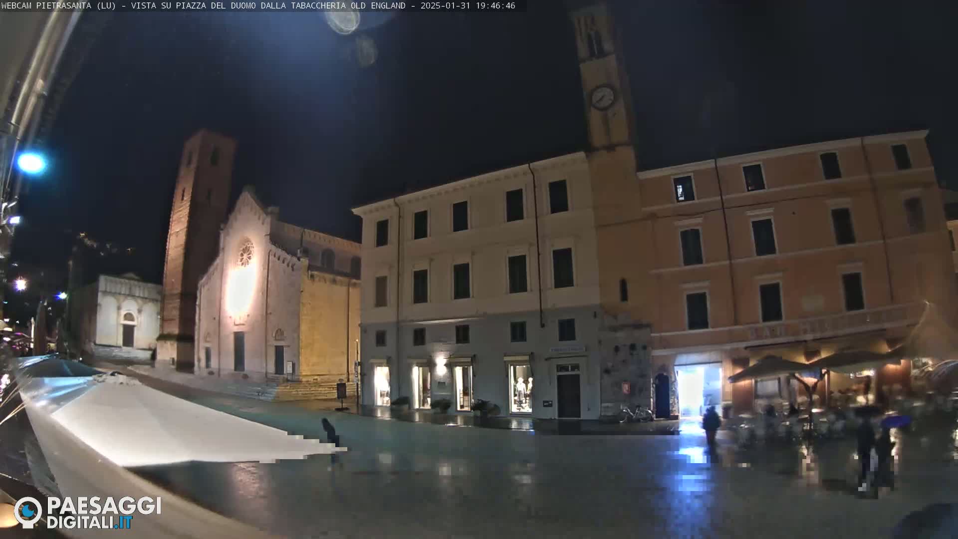A nighttime, rain-slicked town square features a church, several buildings, and a few people walking under the dim light of street lamps.