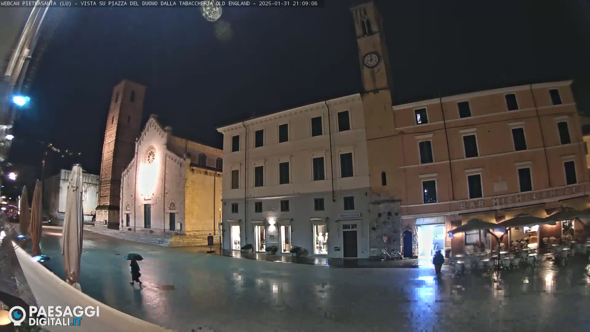 A nighttime, rain-slicked town square features a church, several buildings, and a person with an umbrella walking through the square.