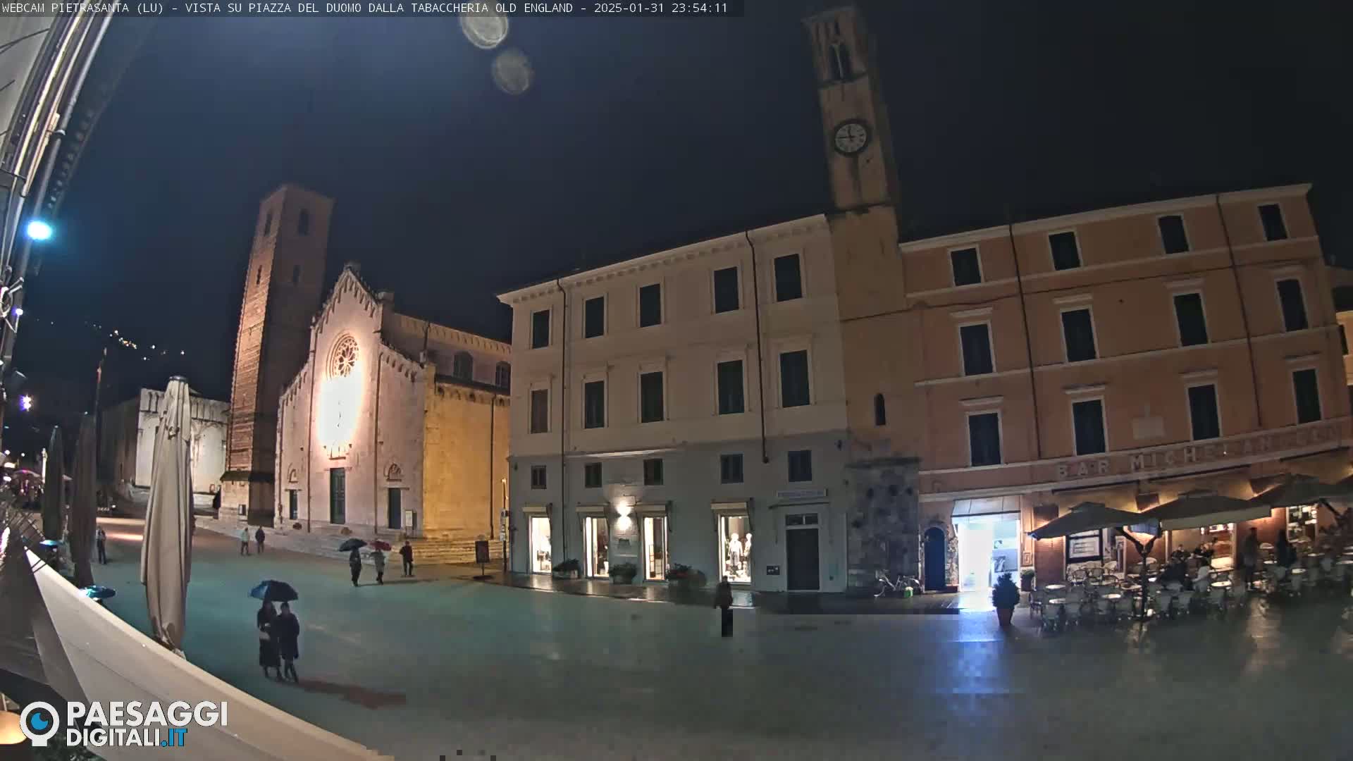 A nighttime view of a town square with a church, several buildings, and a few people walking under umbrellas.