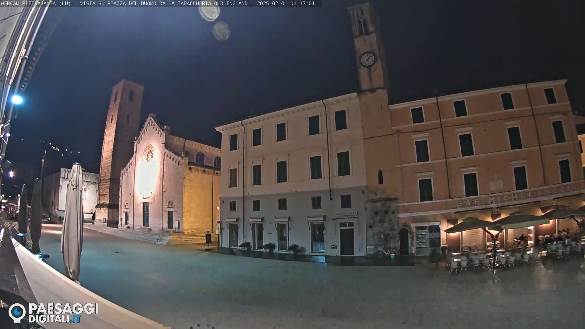 A nighttime view of a town square with several buildings, including a church with a tall bell tower, and an outdoor cafe with umbrellas and tables.