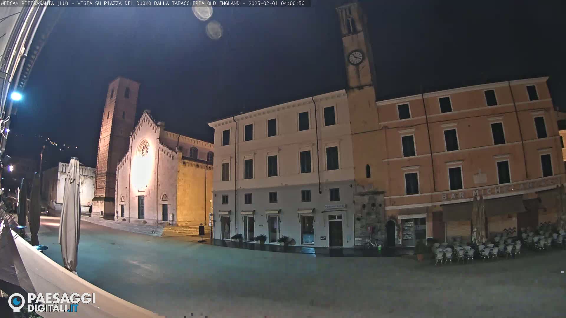 A nighttime view of a town square with several buildings, including a church and a clock tower, under a clear, dark sky.