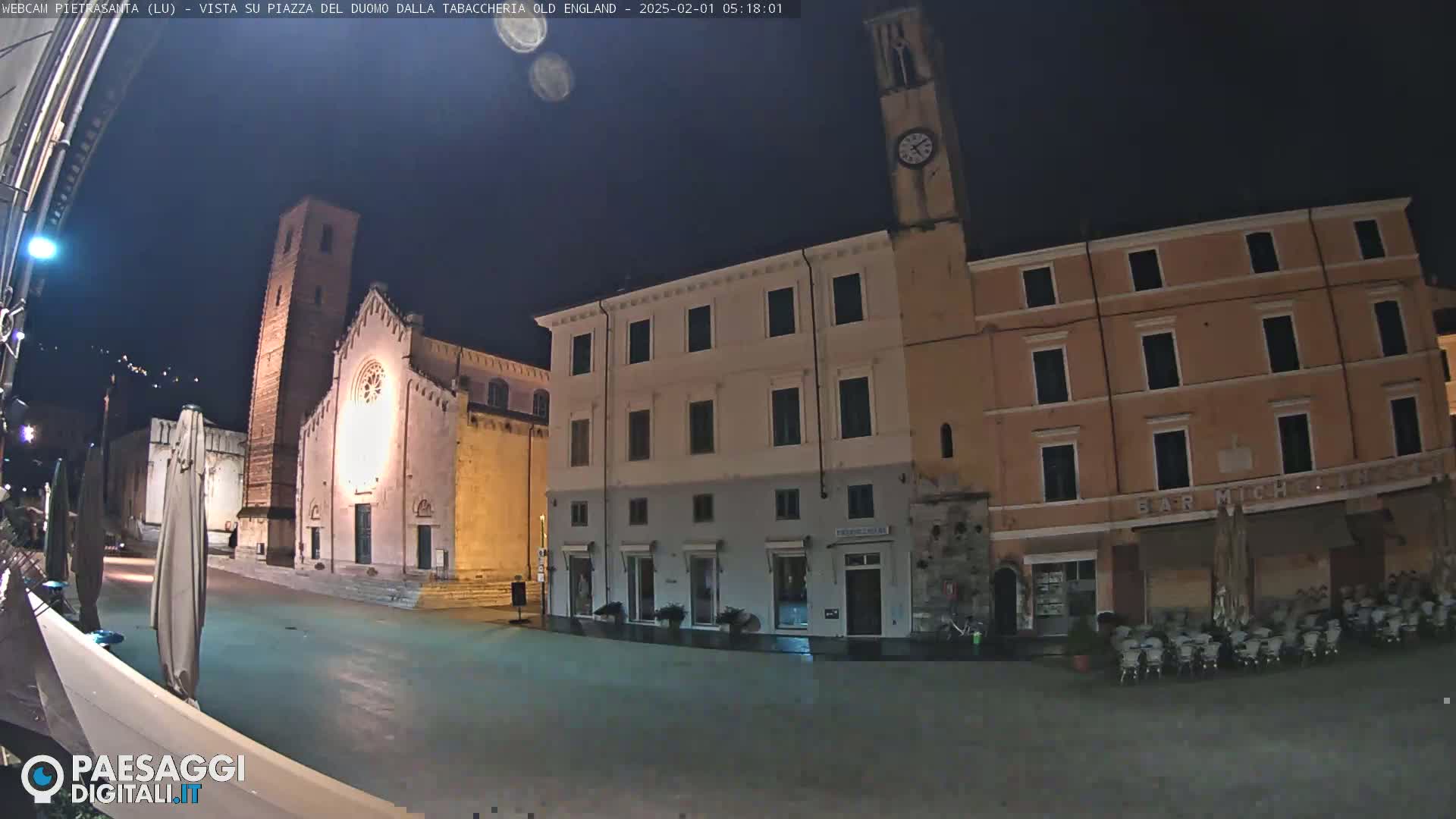 A nighttime view of a town square, showing several buildings including a church with a tall tower and a clock tower, under a clear, dark sky.