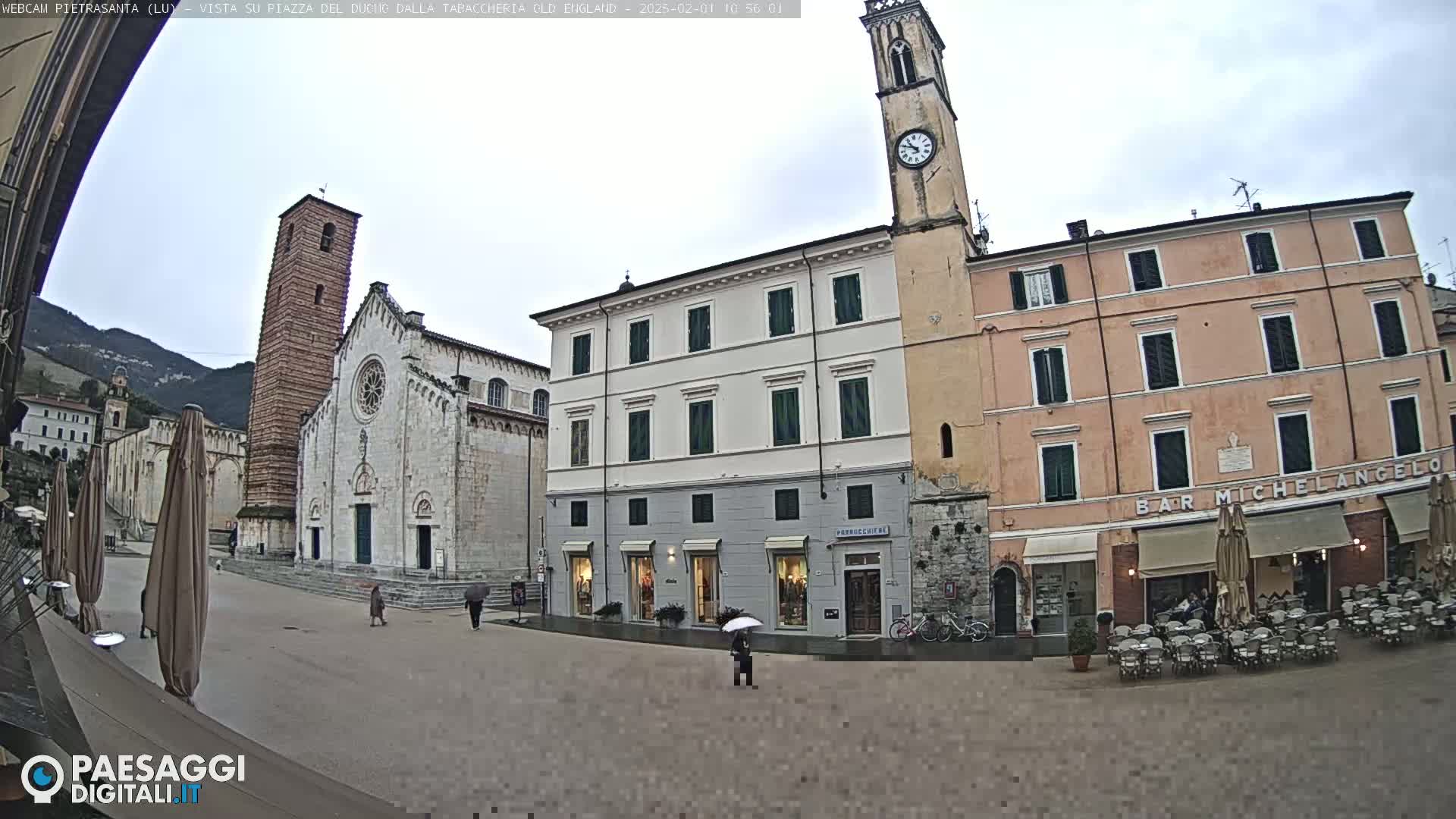 A town square with a church, clock tower, and several buildings under an overcast sky; some people are present, and outdoor seating is set up at a cafe.