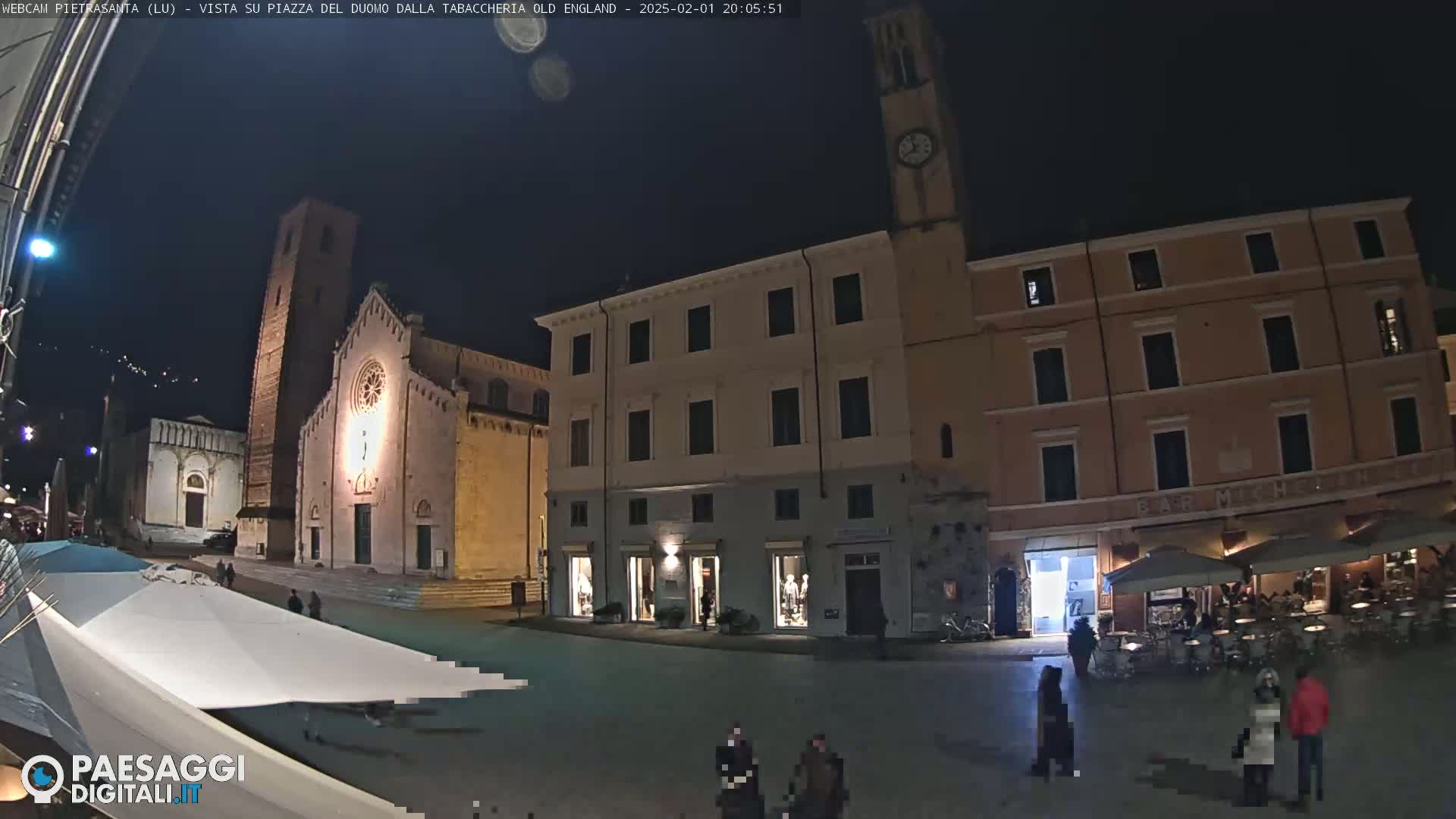 A nighttime view of a town square with a church, buildings, and a few people walking around under a clear, dark sky.