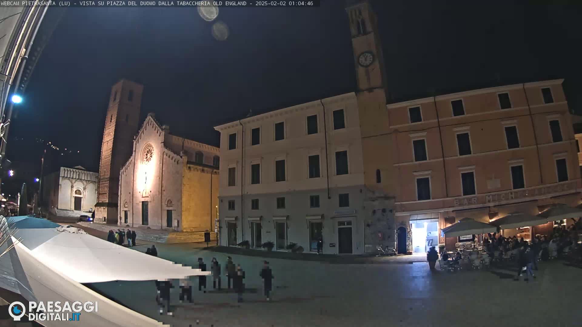 A nighttime view of a town square with a church, buildings, and people gathered in the square under a dark sky.