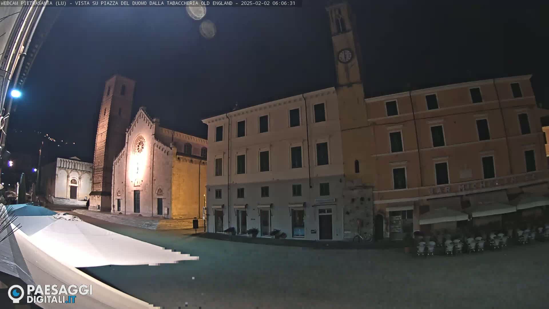 A nighttime view of a town square with a church, several buildings, and outdoor seating under umbrellas.