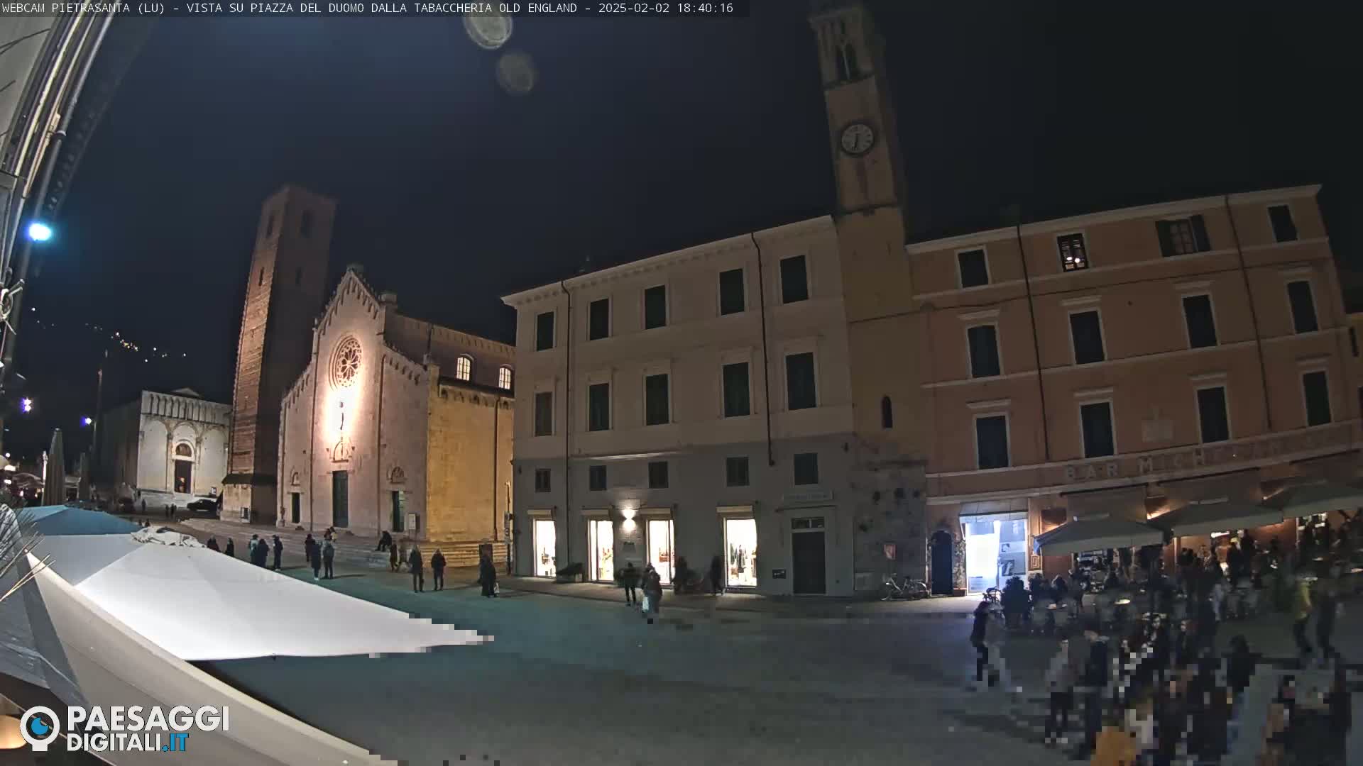 A nighttime view of a town square with a church and several buildings, under a clear, dark sky, showing people gathered in the square and around outdoor seating areas.