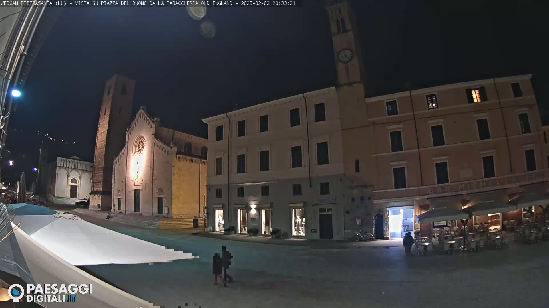A nighttime, outdoor view of a town square with several buildings, including a church with a tall tower, and a cafe with outdoor seating.